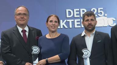 Auf dem Bild zu sehen: Thomas Krull, Annemarie Krull und Daniel Karstens stehen bei der XY-Preisverleihung in Berlin nebeneinander vor einer blauen Wand mit großen, weißen Schriftzügen. 