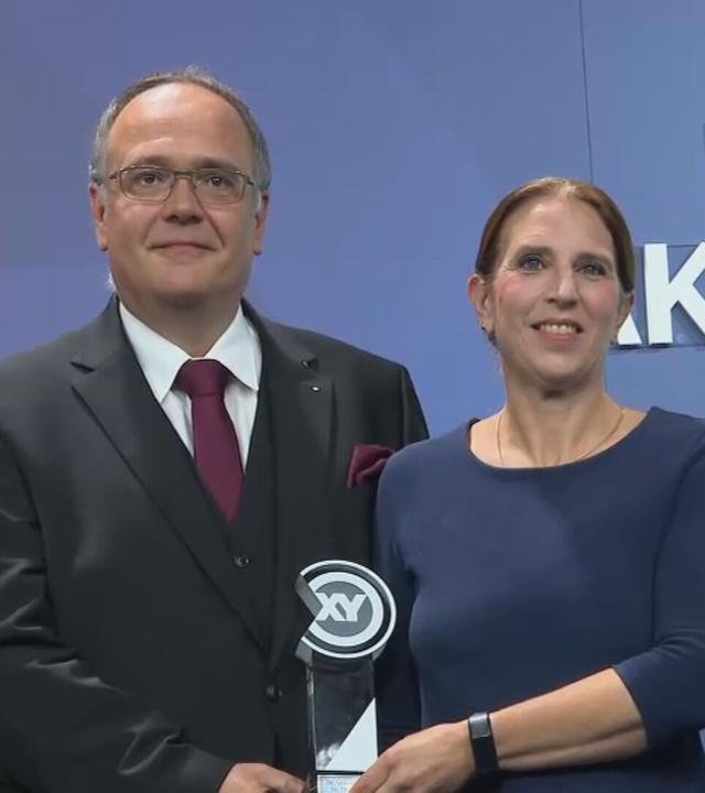 Auf dem Bild zu sehen: Thomas Krull, Annemarie Krull und Daniel Karstens stehen bei der XY-Preisverleihung in Berlin nebeneinander vor einer blauen Wand mit großen, weißen Schriftzügen. 