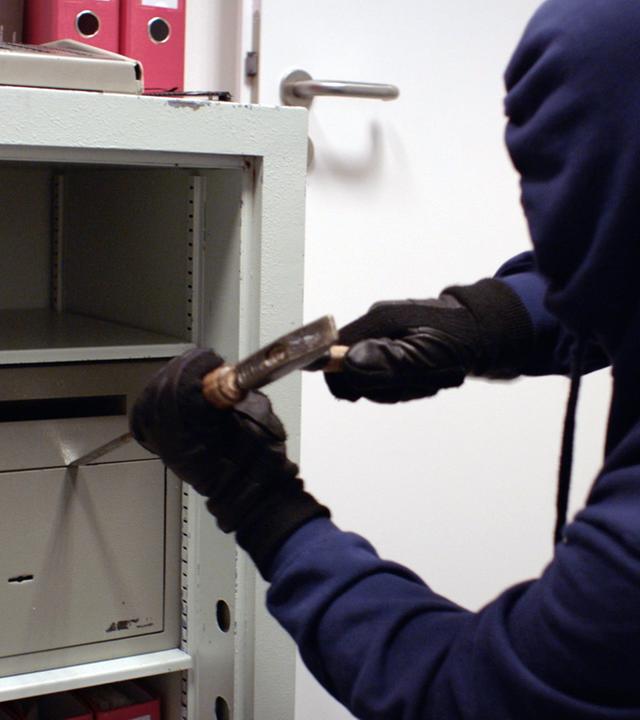 Nachgestellte Szene: Ein dunkel gekleideter Mann mit Kapuze versucht mit Hammer und Meißel einen Tresor zu öffnen.