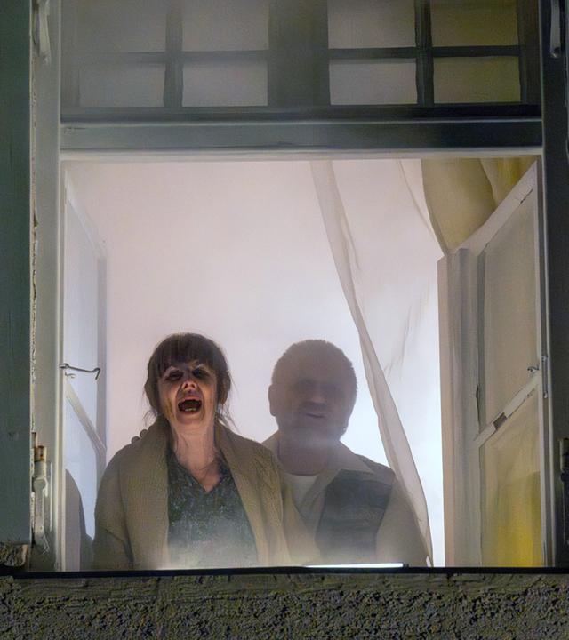 Nachgestellte Szene: Ein älteres Ehepaar steht am Fenster und ruft um Hilfe, während um sie herum das Haus brennt.
