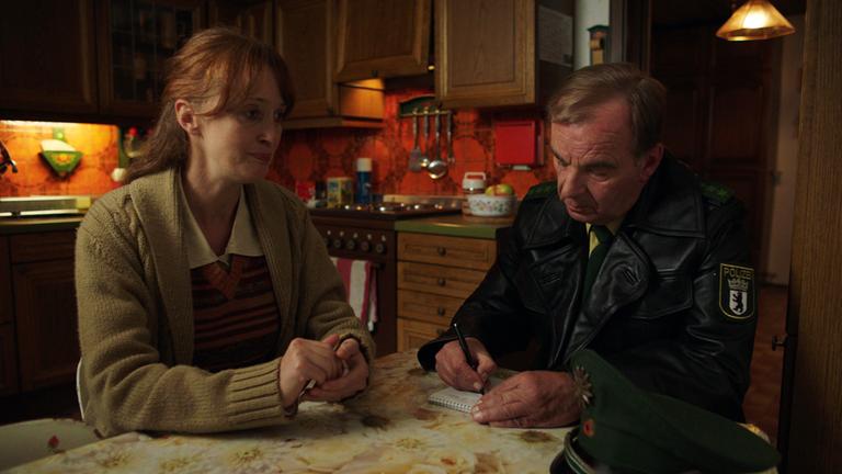 Nachgestellte Szene: Eine Frau und ein Polizist sitzen am Küchentisch. Der Polizist schreibt in einen Notizblock.