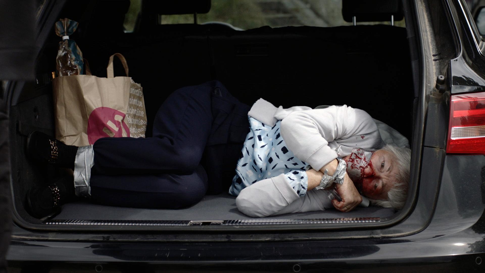 Nachgestellte Szene: Eine Frau liegt im Kofferraum eines schwarzen Audis neben Einkaufstüten. Sie ist im Gesicht blutverschmiert und mit Panzertape gefesselt und geknebelt.