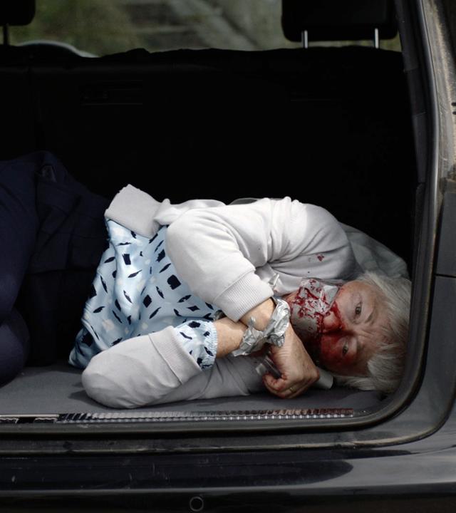 Nachgestellte Szene: Eine Frau liegt im Kofferraum eines schwarzen Audis neben Einkaufstüten. Sie ist im Gesicht blutverschmiert und mit Panzertape gefesselt und geknebelt.