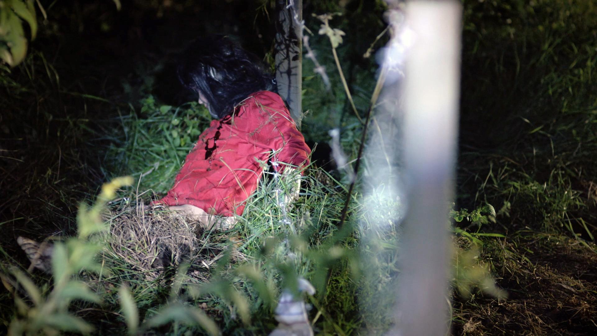 Nachgestellte Szene: Ein kleines dunkelhaariges Mädchen mit rotem Pulli lehnt am Pfosten eines Weidezauns. Ihr Blick ist nach unten gerichtet. Sie ist tot.