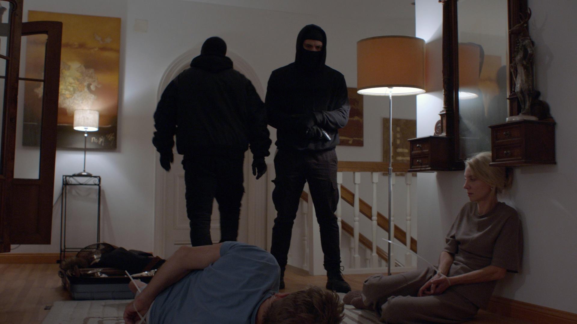 Nachgestellte Szene: Im Flur eines Wohnhauses im Obergeschoss stehen zwei schwarz maskierte Männer, einer davon mit Pistole. Der Familienvater liegt mit Kabelbindern gefesselt am Boden, neben ihm kauert seine Frau, ebenfalls gefesselt.