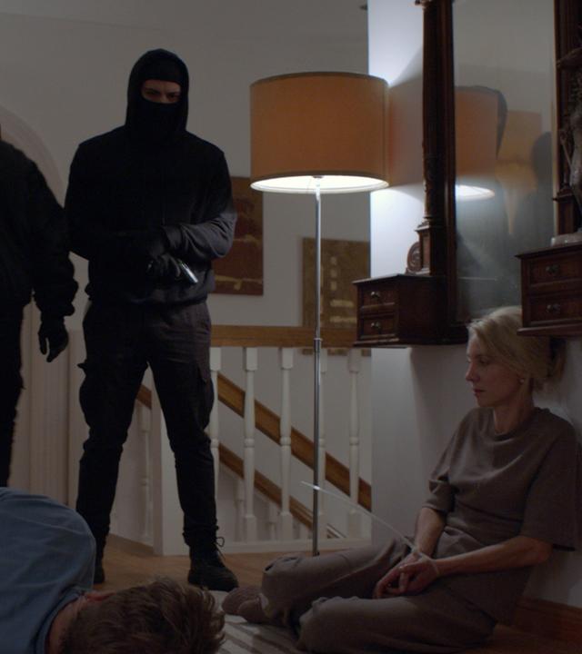 Nachgestellte Szene: Im Flur eines Wohnhauses im Obergeschoss stehen zwei schwarz maskierte Männer, einer davon mit Pistole. Der Familienvater liegt mit Kabelbindern gefesselt am Boden, neben ihm kauert seine Frau, ebenfalls gefesselt.