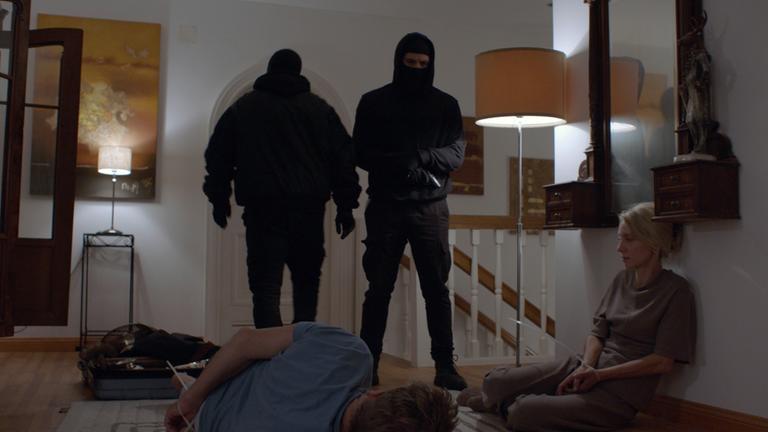 Nachgestellte Szene: Im Flur eines Wohnhauses im Obergeschoss stehen zwei schwarz maskierte Männer, einer davon mit Pistole. Der Familienvater liegt mit Kabelbindern gefesselt am Boden, neben ihm kauert seine Frau, ebenfalls gefesselt.