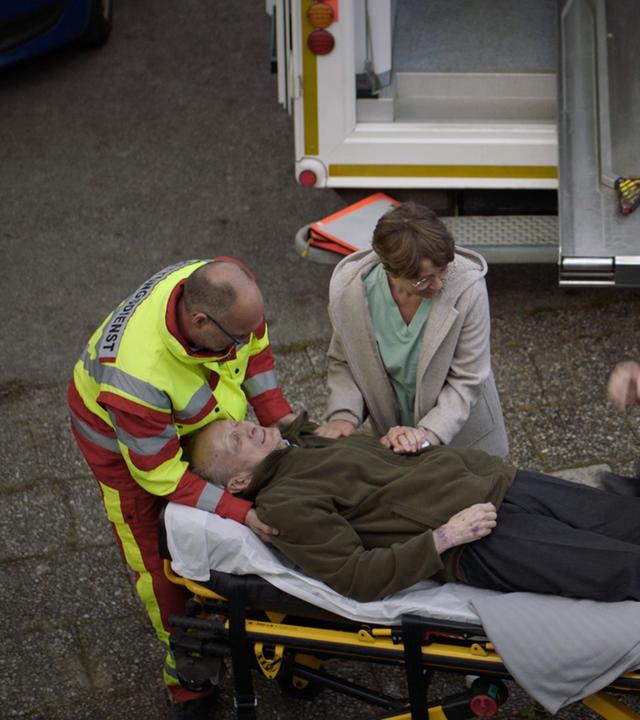 Nachgestellte Szene: Ein alter Mann liegt auf der Liege eines Krankenwagens. Zwei Sanitäter kümmern sich um ihn. Seine Enkelin hält ihm die Hand. Im Hintergrund eine Polizistin.