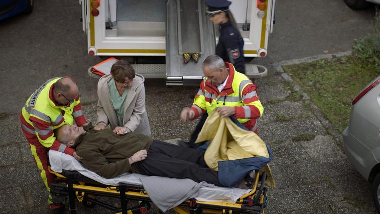 Nachgestellte Szene: Ein alter Mann liegt auf der Liege eines Krankenwagens. Zwei Sanitäter kümmern sich um ihn. Seine Enkelin hält ihm die Hand. Im Hintergrund eine Polizistin.