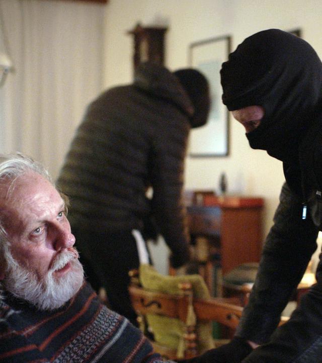 Nachgestellte Szene: Ein schwarz gekleideter Räuber bedroht einen alten Mann. Im Hintergrund durchsucht ein weiterer Täter einen Schrank.