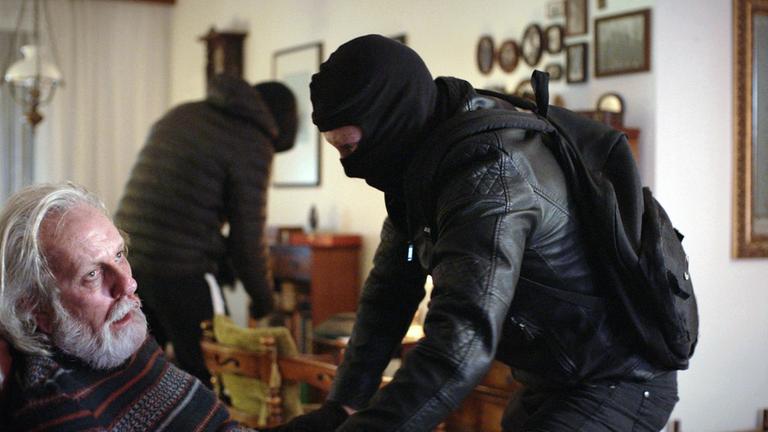 Nachgestellte Szene: Ein schwarz gekleideter Räuber bedroht einen alten Mann. Im Hintergrund durchsucht ein weiterer Täter einen Schrank.