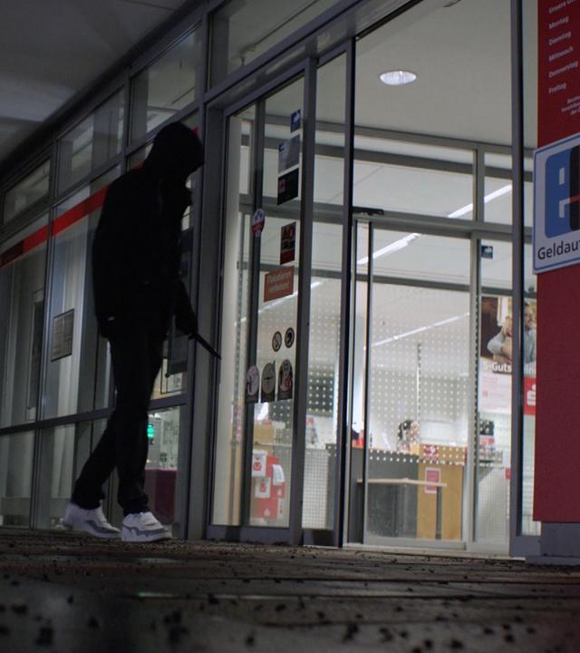 Nachgestelle Szene: Ein vermummter Mann tritt in eine Sparkassenfiliale ein. Er hält ein Messer in der Hand.