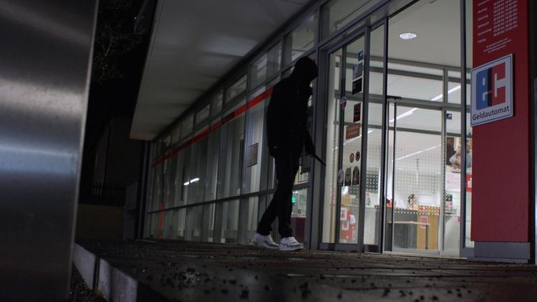 Nachgestelle Szene: Ein vermummter Mann tritt in eine Sparkassenfiliale ein. Er hält ein Messer in der Hand.