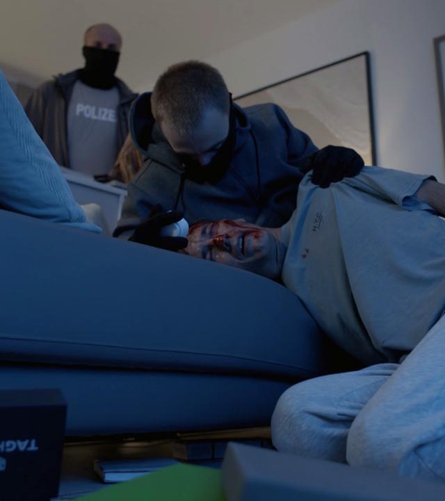 Nachgestellte Szene: Ein Mann liegt blutüberströmt auf einer Couch. Hinter ihm stehen zwei weitere Männer, einer davon fasst ihn grob an der Schulter. Auf dem Boden liegen viele Gegenstände verstreut, darunter eine Schachtel mit der Aufschrift "Tag Heuer"