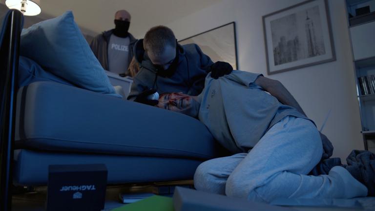 Nachgestellte Szene: Ein Mann liegt blutüberströmt auf einer Couch. Hinter ihm stehen zwei weitere Männer, einer davon fasst ihn grob an der Schulter. Auf dem Boden liegen viele Gegenstände verstreut, darunter eine Schachtel mit der Aufschrift "Tag Heuer"