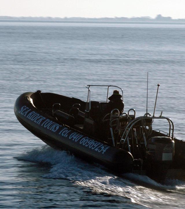 Nachgestellte Szene: Ein Schlauchboot mit Außenbordmotor und einem Mann am Steuerrad fährt Richtung offener See.