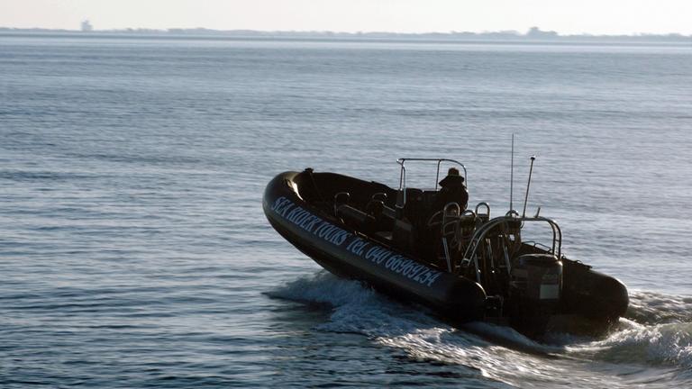 Nachgestellte Szene: Ein Schlauchboot mit Außenbordmotor und einem Mann am Steuerrad fährt Richtung offener See.