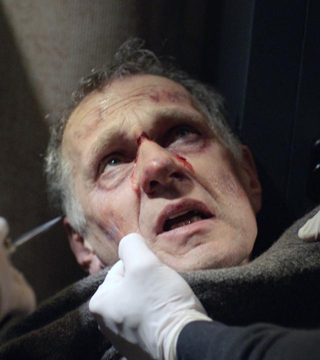 Nachgestellte Szene: Ein blutender Mann wird von einem Unbekannten am Kragen gepackt. Ein zweiter Unbekannter hält ihm ein Messer vors Gesicht.