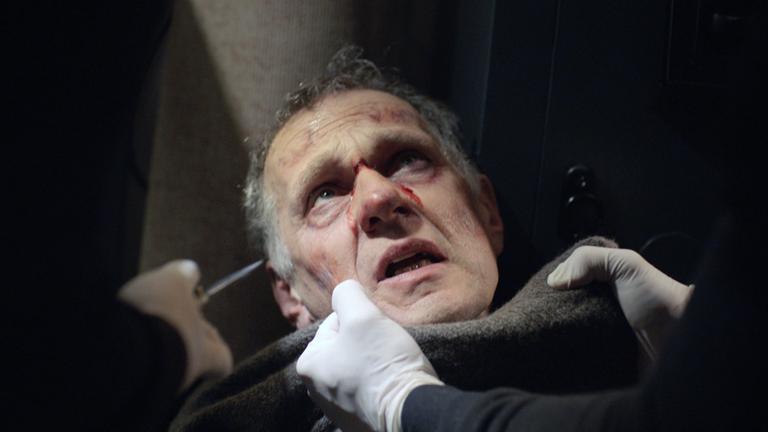 Nachgestellte Szene: Ein blutender Mann wird von einem Unbekannten am Kragen gepackt. Ein zweiter Unbekannter hält ihm ein Messer vors Gesicht.