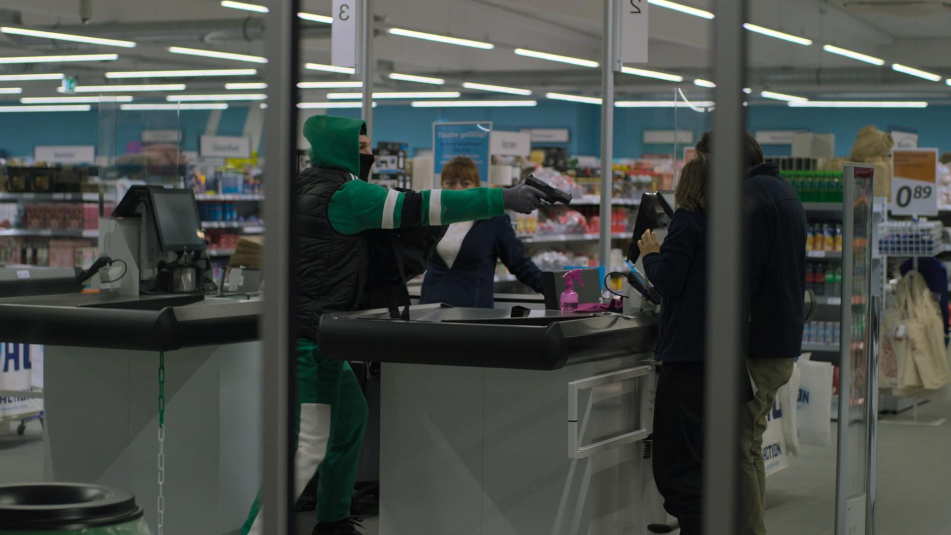 Nachgestellte Szene: Durch die Fensterscheibe eines Discounters ist ein Mann in grünem Jogginganzug und schwarzer Weste zu sehen. Mit gezogener Waffe bedroht er die Angestellten.