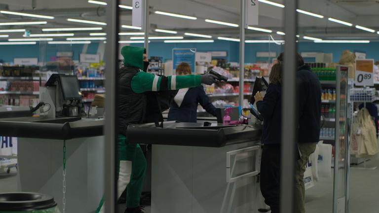 Nachgestellte Szene: Durch die Fensterscheibe eines Discounters ist ein Mann in grünem Jogginganzug und schwarzer Weste zu sehen. Mit gezogener Waffe bedroht er die Angestellten.