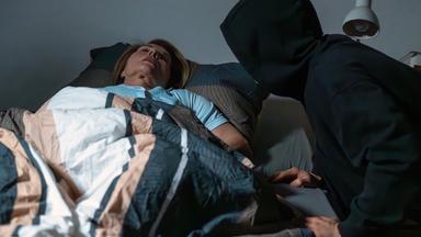 Nachgestellte Szene: Eine ältere Frau liegt schlafend in ihrem Bett. Ein Mann mit schwarzer Kapuze beugt sich über sie.