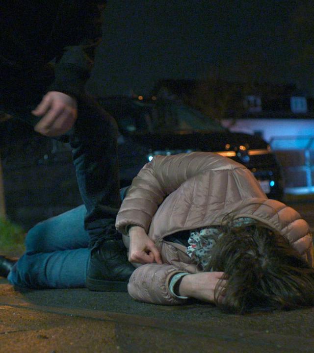Nachgestellte Szene: Eine Frau liegt verletzt auf der Straße. Der Täter steht neben ihr.