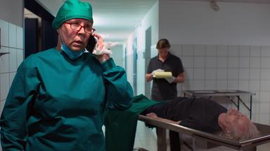 Nachgestellte Szene: Ein lebloser Mann liegt aufgebahrt in der Gerichtsmedizin. Eine Medizinerin telefoniert im Vordergrund.