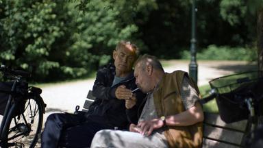 Nachgestellte Szene: Zwei Männer sitzen auf einer Parkbank und rauchen eine Zigarette.