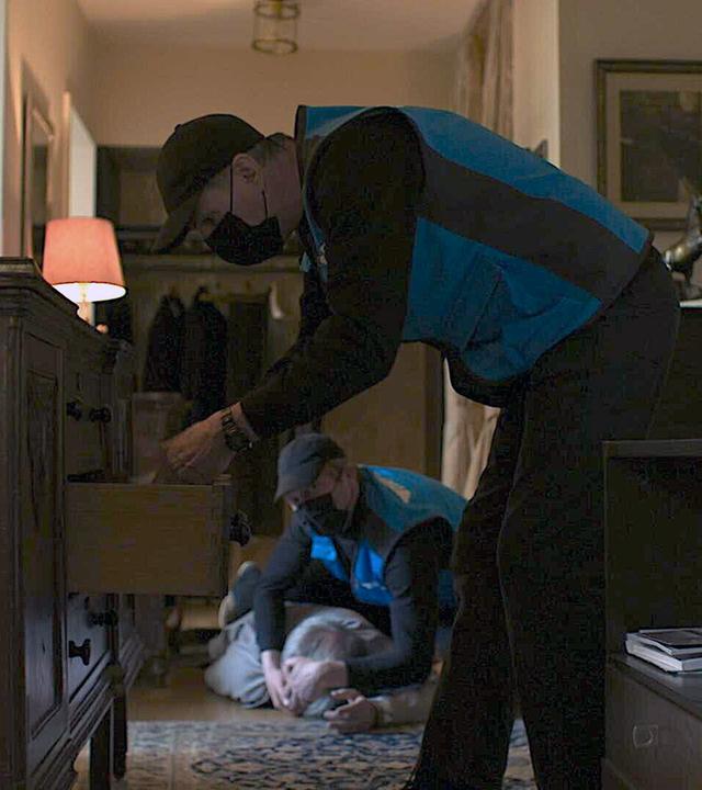 Nachgestellte Szene: Zwei Männer mit Westen eines Paketdienstes in der Wohnung eines älteren Mannes. Einer durchsucht die Schubladen eines Schranks.