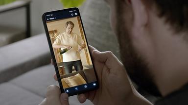 XY Szenenfoto: Ein Mann schaut sich auf dem Handy einen Beitrag eines vermeintlich erfolgreichen Online-Coaches an, der sich ein Glas Sekt einschenkt. 
