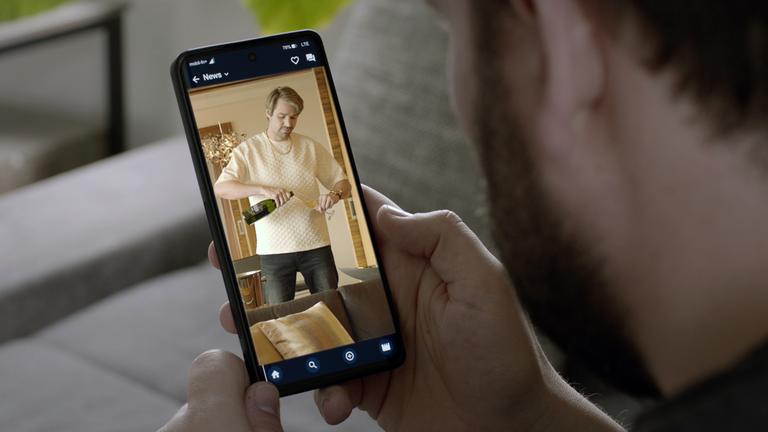 XY Szenenfoto: Ein Mann schaut sich auf dem Handy einen Beitrag eines vermeintlich erfolgreichen Online-Coaches an, der sich ein Glas Sekt einschenkt. 