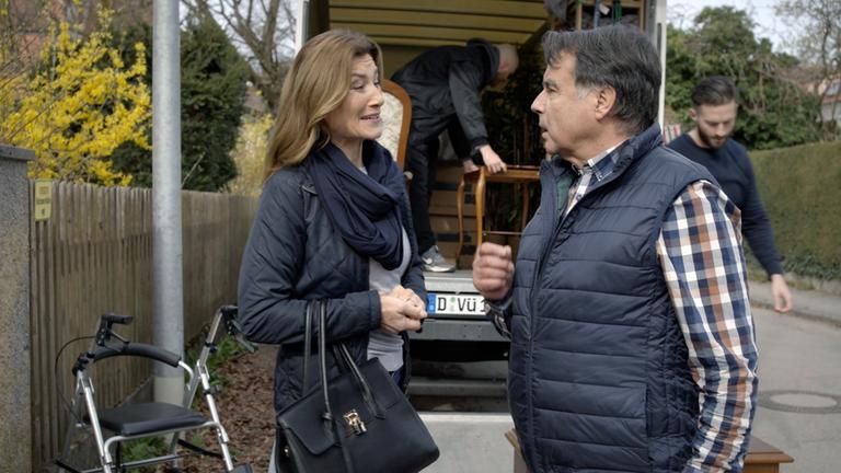 XY-Szenenfoto: Eine Pflegerin und der Senior, den sie betreut, stehen vor einem Möbelwagen, der von zwei Männern beladen wird. 