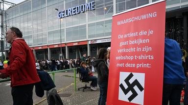Demonstranten stehen neben einem Schild mit der Aufschrift "Achtung, der Künstler, den Sie gleich sehen werden, hat in seinem Webshop T-Shirts mit diesem Aufdruck verkauft: Wir dachten, Sie sollten das wissen", das am GelreDome Stadium vor dem Konzert des Rappers Ye, früher bekannt als Kanye West, am 06.06.2026 aufgebaut ist. 
