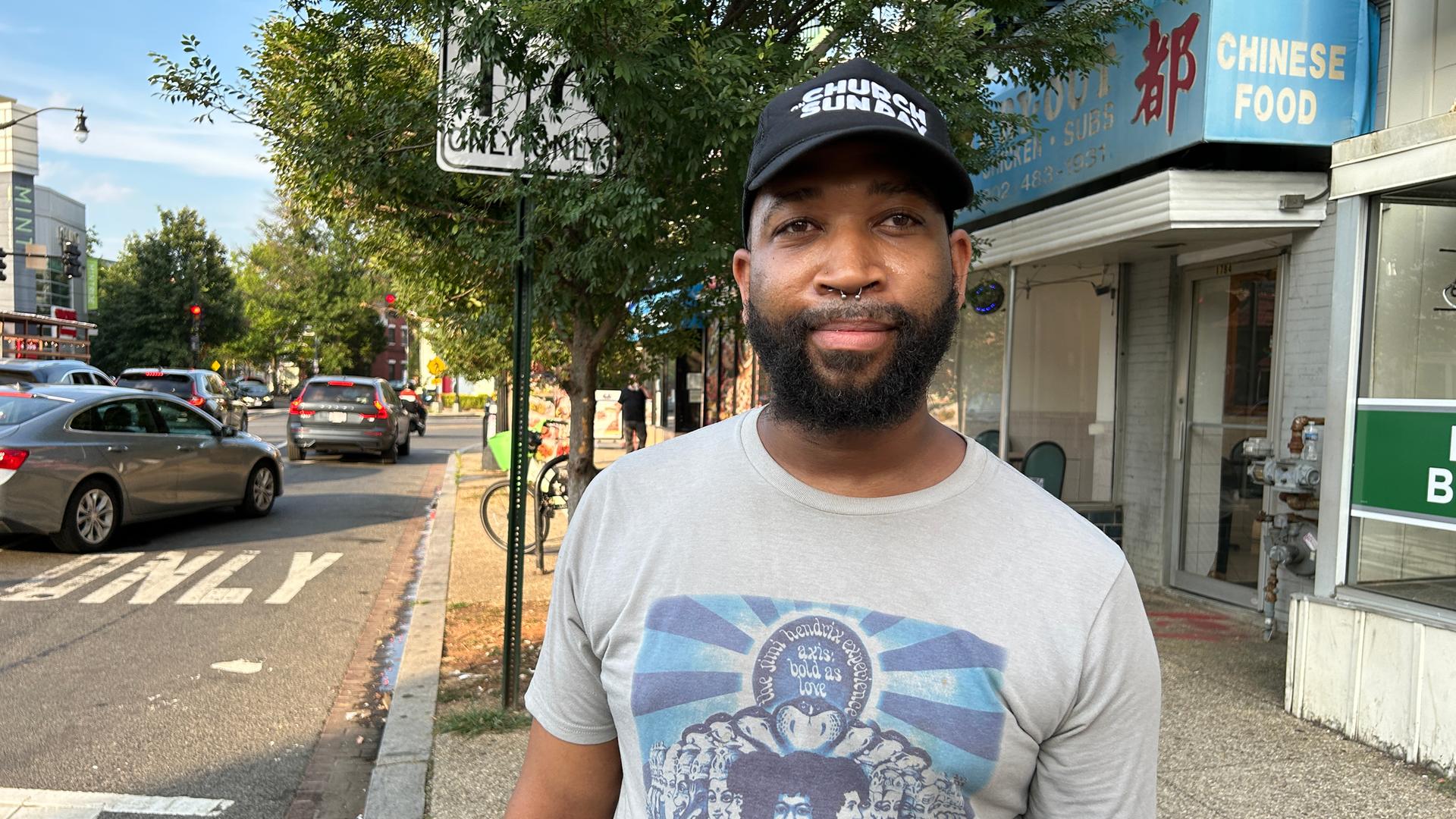 Eine junger schwarzer Mann steht im T-shirt und mit einer Baseballmütze Rand einer Straße.