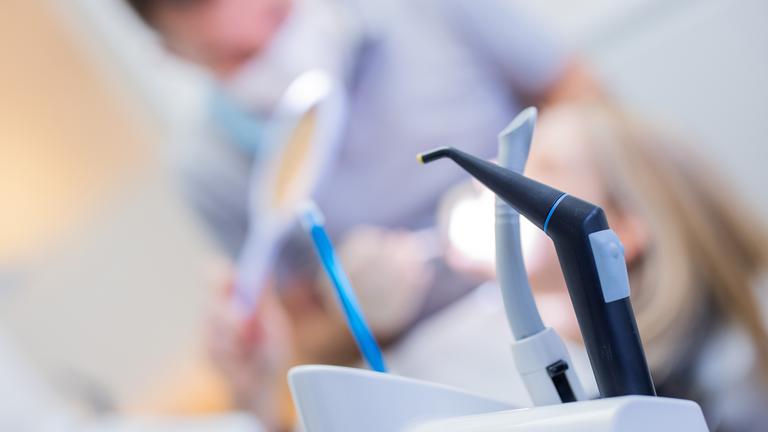 Im Vordergrund sieht man zahnmedizinische Geräte, im Hintergrund eine behandelnde Fachkraft.