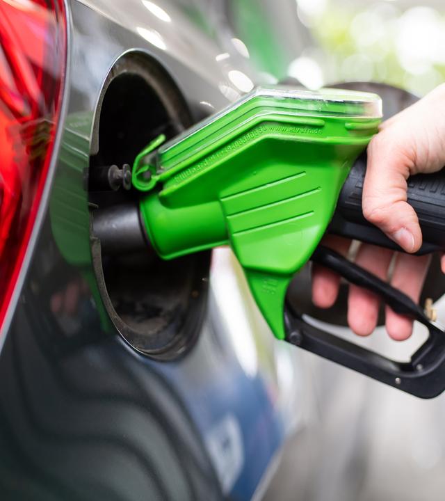 Eine Frau hält an einer Tankstelle an einer Zapfsäule eine Zapfpistole in der Hand und betankt ein Auto.