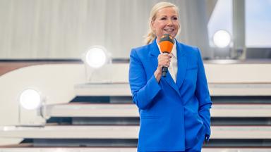 Andrea Kiewel steht auf einer Bühne vor mehreren Stufen und hält ein orangefarbenes Mikrofon.