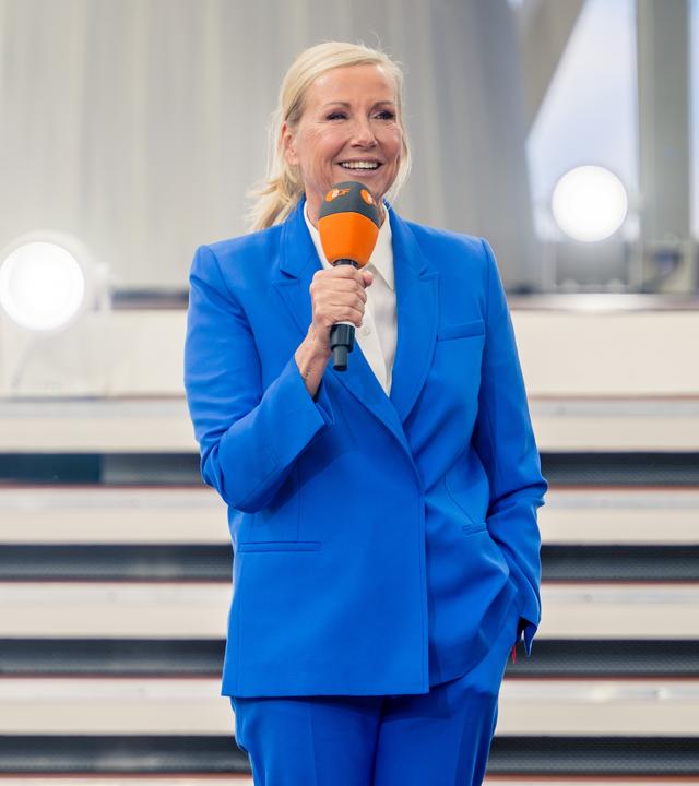 Andrea Kiewel steht auf einer Bühne vor mehreren Stufen und hält ein orangefarbenes Mikrofon.