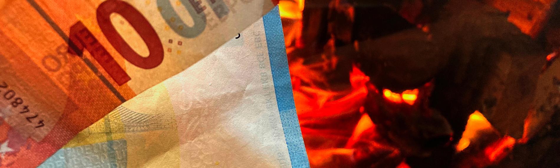 Ein Zehneuroschein und ein Zwanzigeuroschein ragen aus der linken Bildecker hervor. Im Hintergrund auf der rechten Seite sieht man ein offenes Feuer im Kamin mit brennenden Holzscheiten.