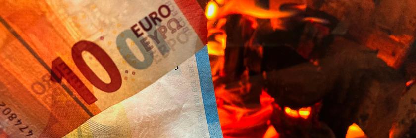 Ein Zehneuroschein und ein Zwanzigeuroschein ragen aus der linken Bildecker hervor. Im Hintergrund auf der rechten Seite sieht man ein offenes Feuer im Kamin mit brennenden Holzscheiten.