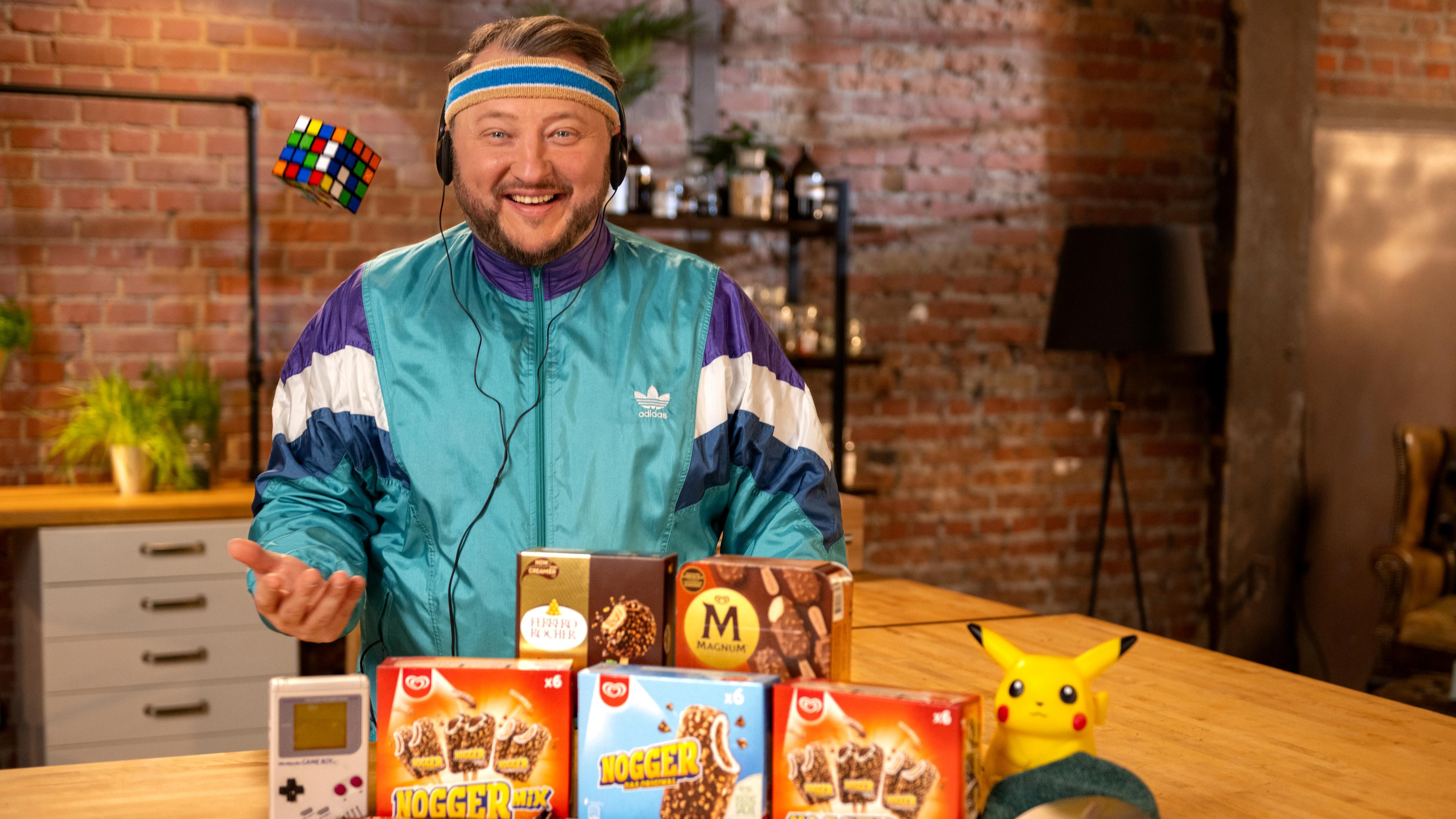 Sebastian Lege schaut freudig in die Kamera. Er hat einen Kopfhörer eines Walkman auf. Vor ihm stehen verschiedene Packungen mit Milcheis am Stiel mit Schokoüberzug.