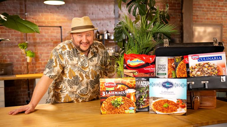 Urlaubsflair aus dem Supermarkt": Sebastian Lege steht, bekleidet mit einem kurzärmligen Hemd und Strohhut, hinter der Küchentheke und lächelt in die Kamera. Vor ihm stehen diverse Packungen mit Lahmacun, einer türkischen Pizza, aus dem Supermarkt rund um zwei Koffer drapiert.