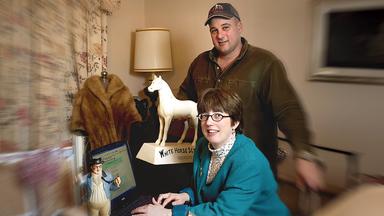 Ina und David Steiner befinden sich in einem Raum, Ina sitzt vor einem Laptop, David steht hinter ihr. Auf dem Tisch steht eine weiße Pferdefigur, an der Seite auf einer Schneiderpuppe hängt eine braune Pelzjacke.