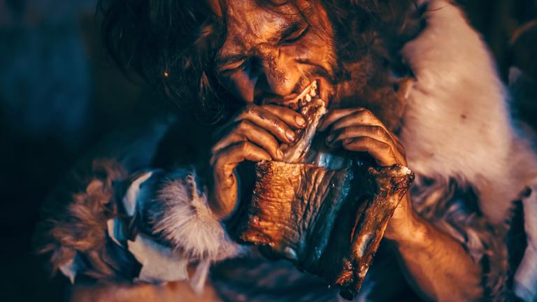 Nahaufnahme, ein Höhlenmensch beißt von einem großen Stück Fleisch ab