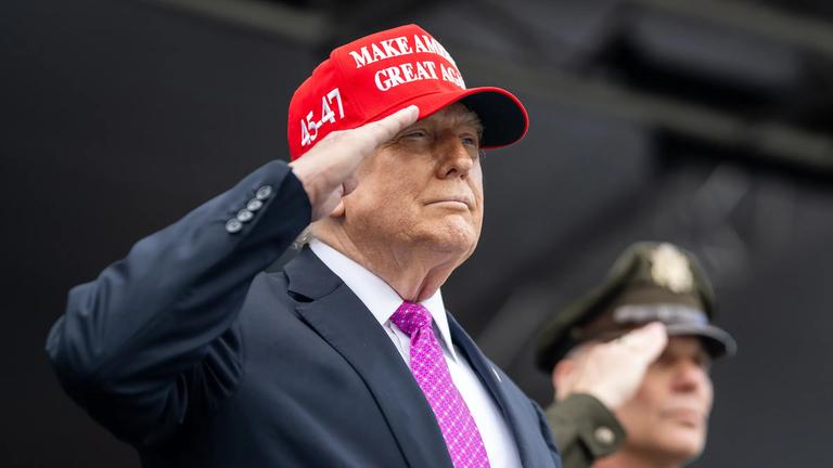 Donald Trump trägt eine rote "Make America Great Again"-Kappe und salutiert. Im Hintergrund steht ein weiterer salutierender Mann, der eine US-amerikanische Militäruniform trägt.