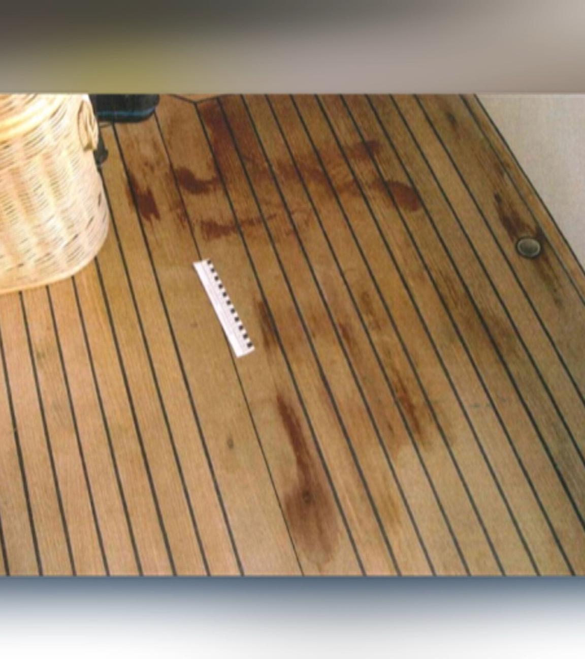 Auf einem braunen Holzboden sind rote Flecken sichtbar. Neben einem beigen Korb liegt ein weiß-schwarzes Lineal, das die Größe der Flecken misst.