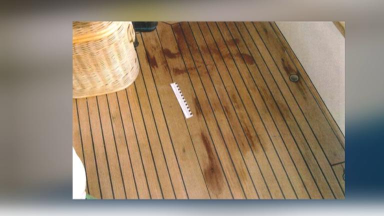 Auf einem braunen Holzboden sind rote Flecken sichtbar. Neben einem beigen Korb liegt ein weiß-schwarzes Lineal, das die Größe der Flecken misst.