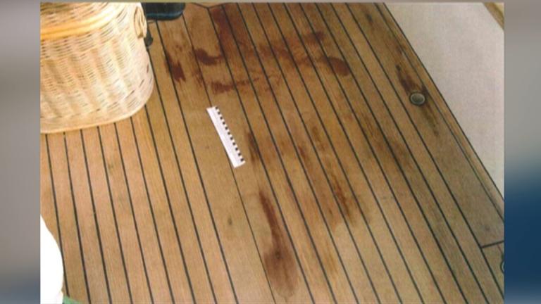 Auf einem braunen Holzboden sind rote Flecken sichtbar. Neben einem beigen Korb liegt ein weiß-schwarzes Lineal, das die Größe der Flecken misst.