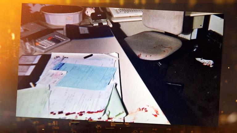 Die Farbfotografie zeigt einen Schreibtisch in einem Büro oder Verwaltungsraum. Im linken Bildbereich liegen mehrere übereinander gestapelte Blätter und Formulare auf dem hellen Tisch – teils weiß, teils blau. Einige der Dokumente sind am unteren Rand deutlich mit blutigen Abdrücken oder Spritzern beschmutzt. Ein schwarzer Kugelschreiber liegt zwischen den Papieren.